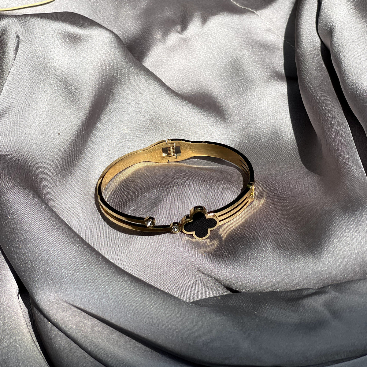 A gold-toned bangle bracelet with a clover motif, resting on draped silver satin fabric.