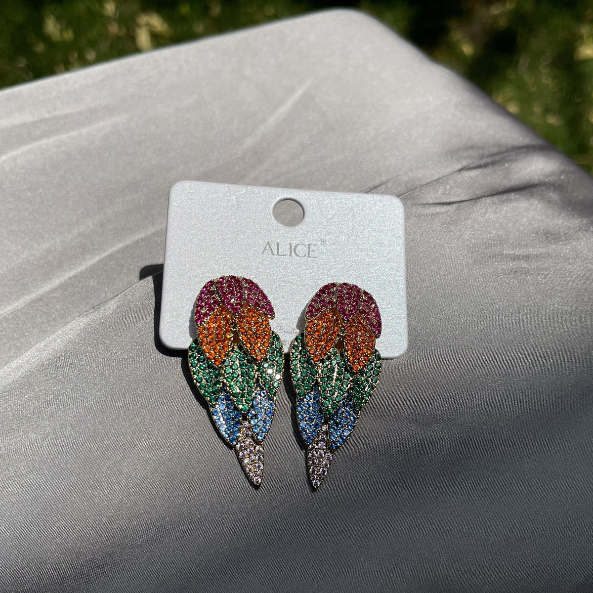 A pair of colorful feather-shaped earrings on a white card labeled ALICE, resting on a gray fabric surface with blurred green grass in the background.