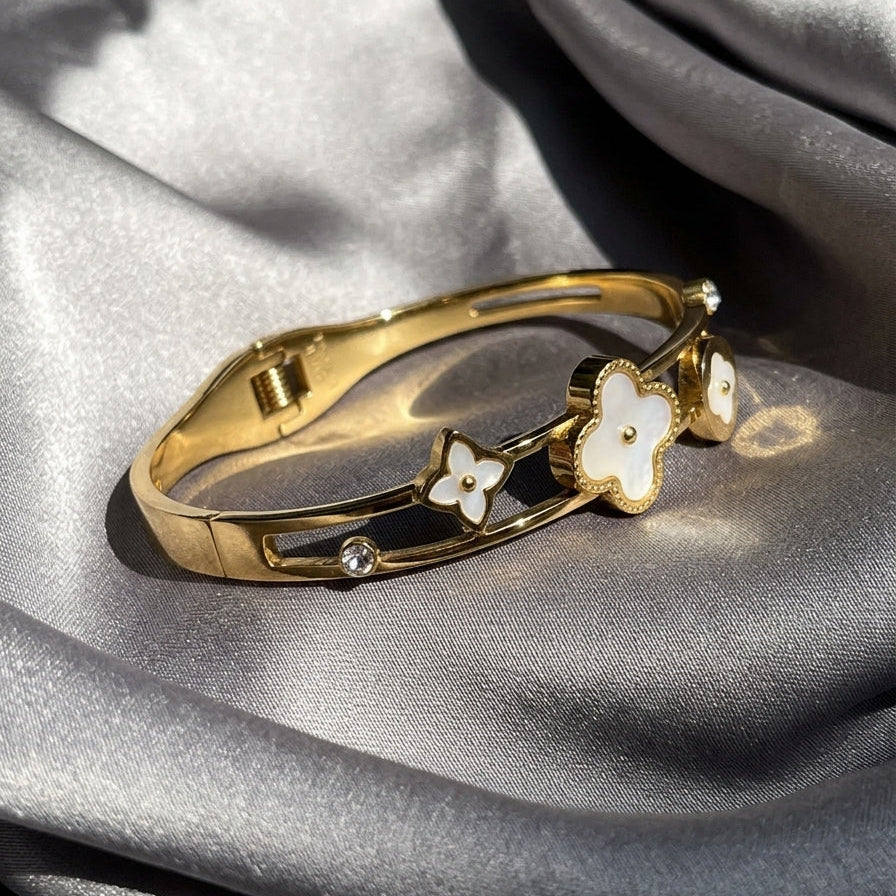 A gold-plated bangle bracelet with white enamel flower charms, resting on draped gray satin fabric.