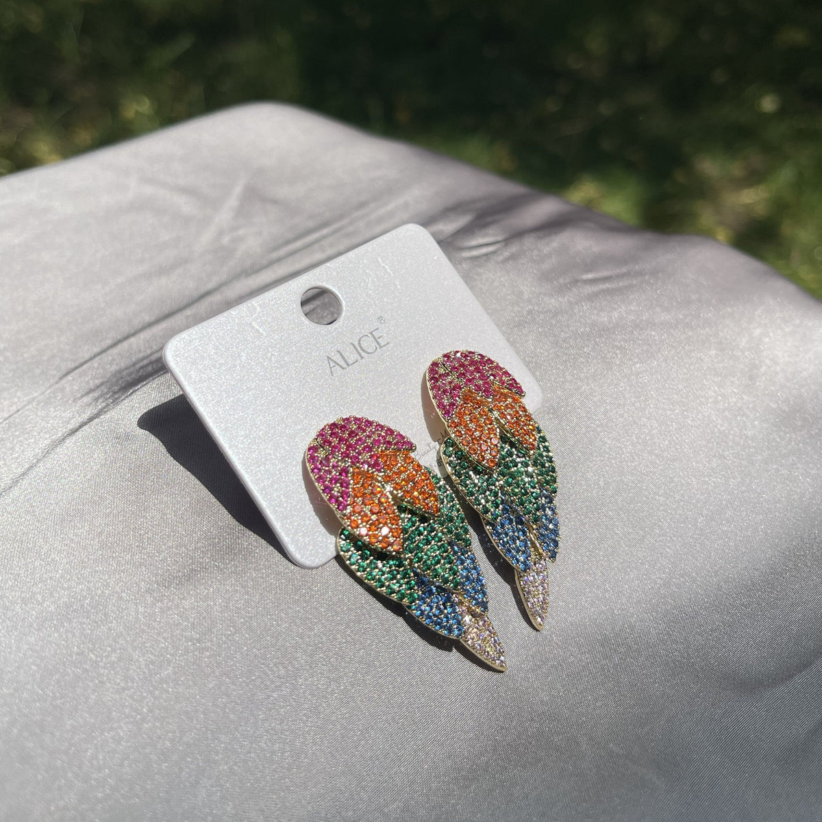A pair of colorful tulip-shaped stud earrings on a white display card labeled ALICE, resting on a gray satin fabric with a blurred green outdoor background.