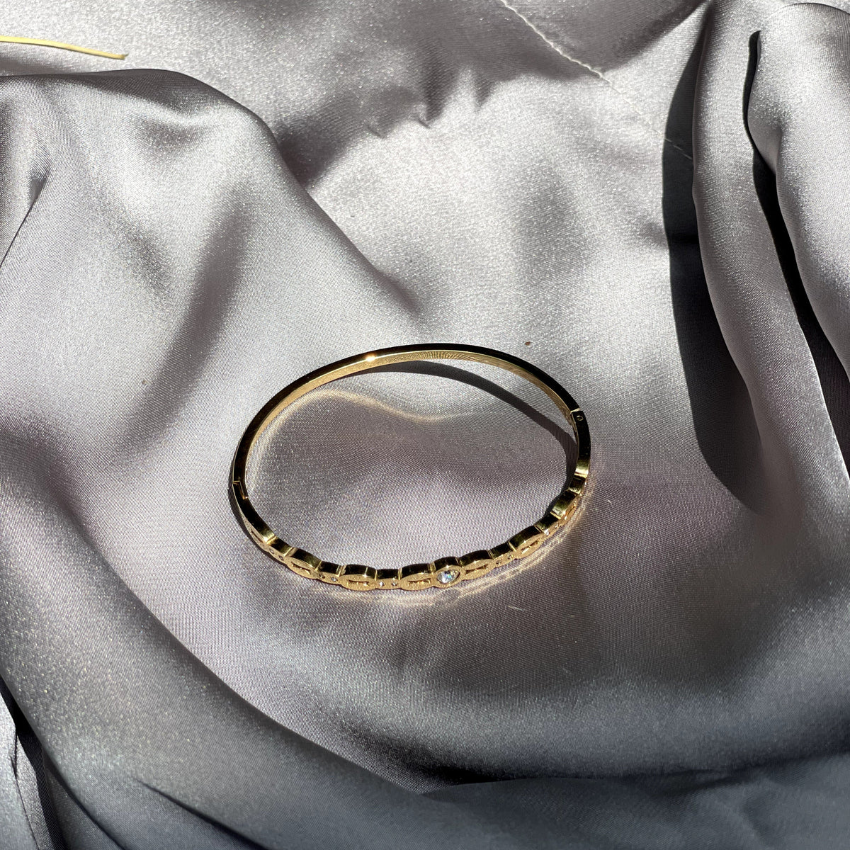 A gold bangle bracelet with a textured, beaded outer edge, displayed on draped silver satin fabric.