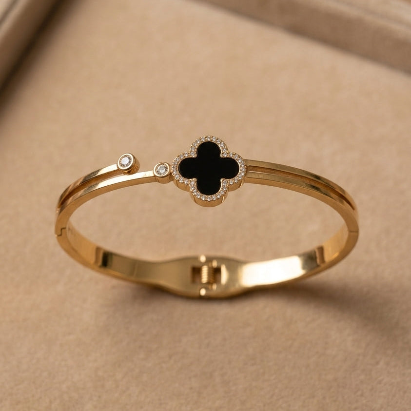 A gold-plated bangle bracelet featuring a central black clover motif surrounded by small clear stones, resting on a beige jewelry display box.