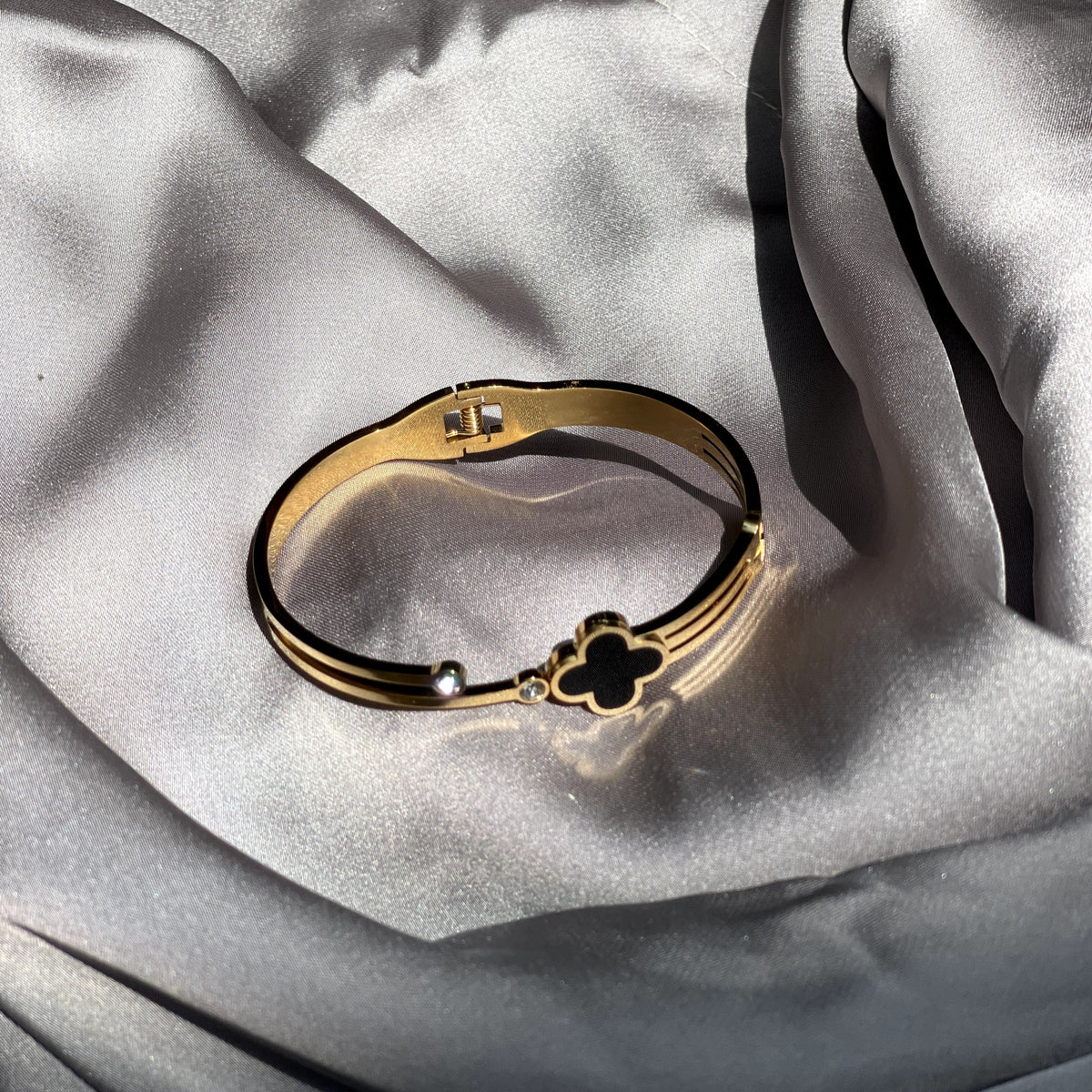 A gold-toned bangle bracelet with a small black clover charm and a tiny clear stone, displayed on draped light gray satin fabric.