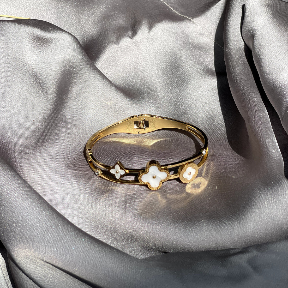 A gold bangle bracelet with three clover motifs, featuring white enamel details, resting on draped gray satin fabric.