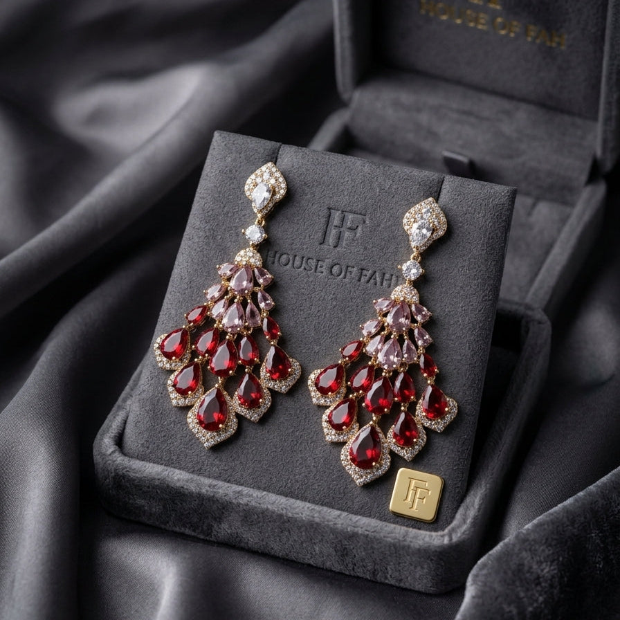 A pair of ornate drop earrings displayed on a velvet jewelry box, set against a draped dark gray fabric background. The earrings feature deep red teardrop-shaped stones and clear crystal accents in a gold-tone setting.