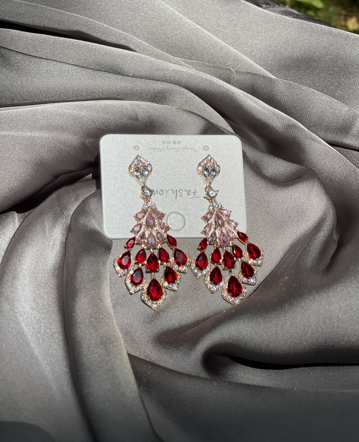 A pair of dangling chandelier earrings with red teardrop crystals and clear rhinestone detailing on a white display card, set against a draped gray satin fabric background.